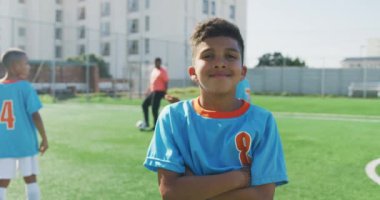 Mavi tişört giyen, yeşil bir futbol sahasında duran ve kameraya bakıp gülümseyen melez bir çocuk portresi.