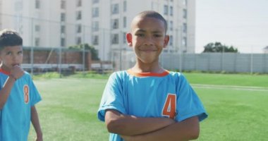 Afrikalı Amerikalı bir çocuğun portresi, futbolcu, mavi tişört giyiyor, yeşil bir futbol sahasında duruyor, kameraya bakıyor ve gülümsüyor.