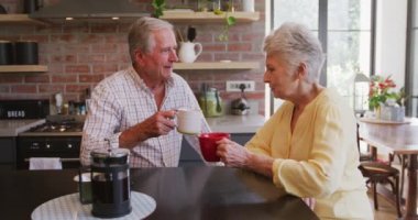 Üst düzey beyaz çift, Coronavirus covid salgını sırasında karantinada karantinada kendi başlarına vakit geçiriyorlar. Masada oturuyorlar, bardaklarını tutuyorlar, ağır çekimde.