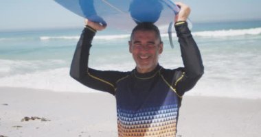 Yaşlı beyaz bir adamın plajda eğlenen portresi, ayakta duran ve başının üstünde sörf tahtası tutan, güneşli bir günde yavaş çekimde kameraya bakan. Rahatlatıcı Yaz Sahil Tatili.