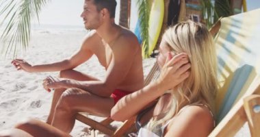 Beyaz bir çift kumsalda eğleniyor, güneşli bir günde bir palmiye ağacının yanında güneşli yataklarda oturuyor ve ağır çekimde gülümsüyor. Rahatlatıcı Yaz Sahil Tatili.