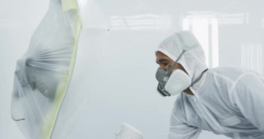 Bir şehir atölyesinde çalışan karışık ırk erkek araba tamircisi koruyucu giysiler giyiyor, sprey tabanca kullanıyor, yavaş çekimde araba boyuyor.