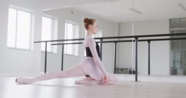 Beyaz kadın balerin, siyah tricot giyiyor, parlak bir stüdyoda dans kursuna hazırlanıyor, yavaş çekimde yere uzanıyor..