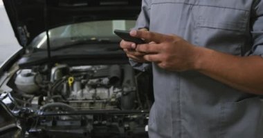 Afrika kökenli Amerikalı erkek araba tamircisi, bir kasaba atölyesinde çalışıyor, açık bir araba motoruna bakıyor, akıllı telefonu yavaş çekte kullanıyor.