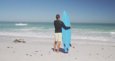 Kıdemli beyaz adam kumsalda eğleniyor, güneşli bir günde ağır çekimde sörf tahtası tutuyor. Rahatlatıcı Yaz Sahil Tatili.