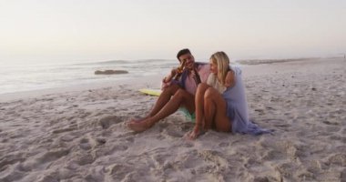Kafkasyalı çift kumsalda vakit geçirmeyi seviyor, gün batımında battaniyeyle kaplanmış, bira şişelerini yavaş çekimde kucaklıyor ve tutuyorlar. Rahatlatıcı Yaz Sahil Tatili.