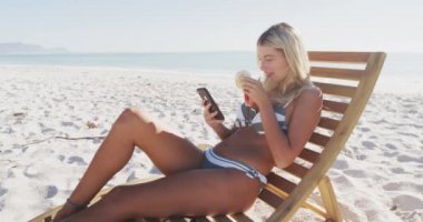 Beyaz bir kadın kumsalda eğleniyor, güneşli bir günde güneşli bir yatakta oturup içki içiyor, akıllı telefonunu yavaş çekimde kullanıyor. Rahatlatıcı Yaz Sahil Tatili.