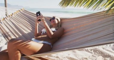 Beyaz bir kadın kumsalda eğleniyor, hamakta uzanıyor ve güneşli bir günde ağır çekimde akıllı telefonunu kullanıyor. Rahatlatıcı Yaz Sahil Tatili.