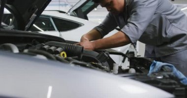 Afro-Amerikalı erkek araba tamircisi, bir kasaba atölyesinde çalışıyor, açık bir araba motoruna bakıyor, bir şeyin yavaş çekte bozulup bozulmadığını kontrol ediyor.