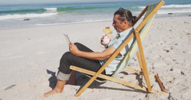 夏のビーチ休暇 ビーチで時間を楽しんでいるシニア白人男性 日向ぼっこに座って 彼のタブレットを使用して スローモーションで晴れた日にドリンクを飲みます — ストック動画