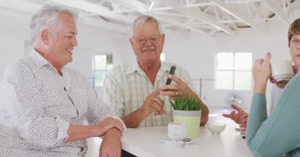 社交ダンス教室の前にテーブルに座り ゆっくりとした動きの中でスマホを使ってお茶やコーヒーを飲みながら話をする幸せな年配の男女の友人たち — ストック動画