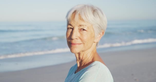 暑假海滩度假 一个年长的高加索女人在海滩上享受时光的画像 看着相机 慢镜头背景下蓝天蓝海的笑容 — 图库视频影像