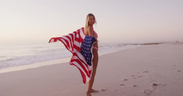 海岸での時間を楽しんでいる白人女性は 遅い動きの日没の間にカメラを見て 米国の旗を握って手を振っています リラックスした夏のビーチ休暇 — ストック動画