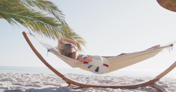 在阳光灿烂的日子里 白人妇女在海滩上享受时光 躺在吊床上 头戴太阳帽 动作缓慢 放宽暑期泳滩假期 — 图库视频影像