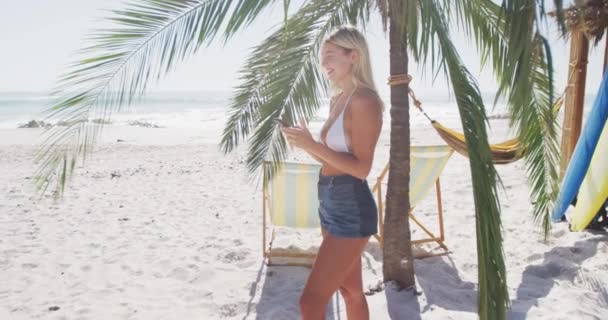 ヤシの木のそばに立って笑顔でビーチで時間を楽しむ白人女性は スローモーションで晴れた日にスマートフォンを使用しています リラックスした夏のビーチ休暇 — ストック動画
