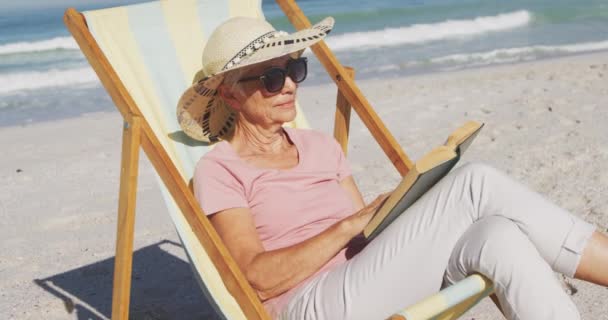 夏のビーチ休暇 ビーチで時間を楽しんでいるシニア白人女性 日向ぼっこに座って スローモーションで晴れた日に本を読む — ストック動画