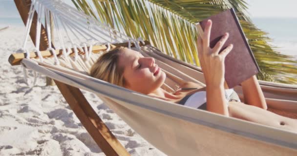 ビーチで時間を楽しんでいる白人女性は ハンモックに横たわって 遅い動きで晴れた日に本を読んでいます リラックスした夏のビーチ休暇 — ストック動画