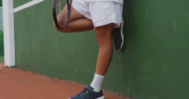Tenis kıyafetleri giyen, tenis raketi tutan, duvara yaslanan, tenis kortunda vakit geçiren beyaz bir adam..