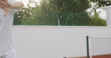Beyazlar içinde tenis oynayan, güneşli bir günde tenis oynayan, tenis raketi tutan, tenis topuna yavaş çekimde vuran, beyaz tenli bir adamın orta bölümü..