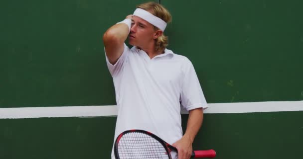 テニスの白人を着た白人男性がコートで時間を過ごし テニスラケットを持ち 壁にもたれて スローモーションで — ストック動画