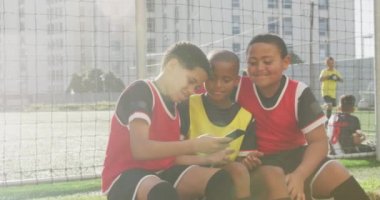 Çoklu etnik gruptan futbol oyuncuları takım elbiselerini giyip oyun sahasının kenarında oturuyorlar, akıllı telefonları güneşte bir arada tutuyorlar ve selfie çekiyorlar.