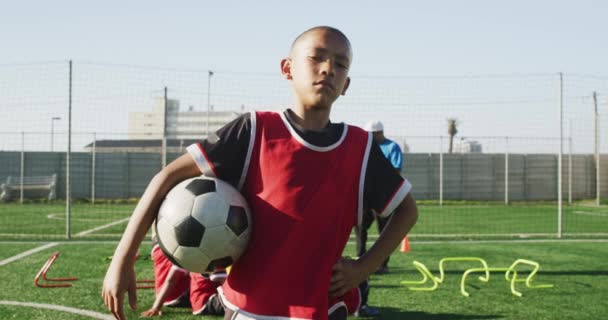 晴れた日に競技場に立つアフリカ系アメリカ人の少年サッカー選手の肖像 トレーニング中に休息を取り カメラを見て サッカーを開催し 背景にチームメイトと一緒に — ストック動画