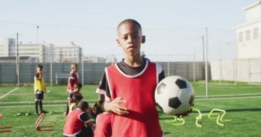 Güneşli bir günde bir oyun sahasında durup antrenman sırasında dinlenen, elinde futbol topu tutan ve arka planda takım arkadaşları olan Afrikalı Amerikalı bir futbolcunun portresi.