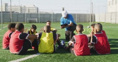 Karışık ırk erkek futbol koçu, futbol antrenmanı sırasında çok ırklı erkek futbol oyuncularından oluşan bir gruba güneşin altında bir oyun alanında talimat veriyor.