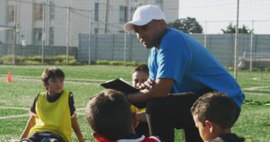 Karışık ırk erkek futbol koçu, futbol antrenmanı sırasında çok ırklı erkek futbol oyuncularından oluşan bir gruba güneşin altında bir oyun alanında talimat veriyor.