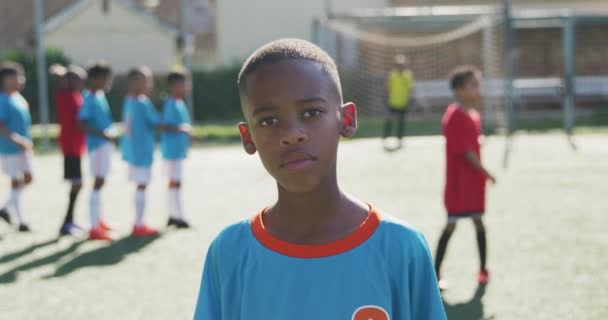 青いTシャツを着て緑のサッカーピッチに立ってカメラを見て笑っているアフリカ系アメリカ人の少年の肖像画 — ストック動画