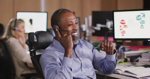 現代的なオフィスで夜遅くに働いている混合レースのビジネスマンは 電話のヘッドセットの話を着て机に座って スローモーションで笑顔 — ストック動画