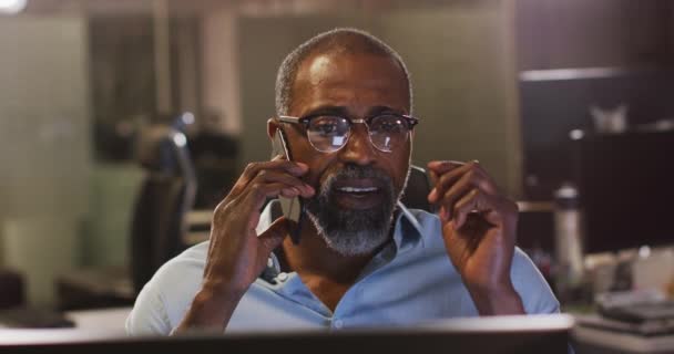 混合种族商人深夜在一间现代化的办公室工作 坐在桌子旁 用智能手机说话 慢吞吞地摘下他的阅读眼镜 — 图库视频影像