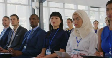 Bir iş seminerinde izleyicilerin önünde oturan çok ırklı iş kadını ve iş adamlarından oluşan çok yönlü giyinmiş bir grup yavaş çekimde sunumu dikkatle dinliyor.