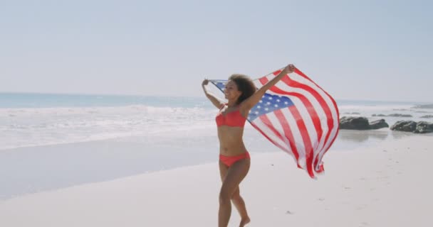 在阳光明媚的日子里 假日里 穿着比基尼在海滩上跑步和跳舞 举着美国国旗 微笑着 背景是大海和蓝天 动作缓慢 — 图库视频影像