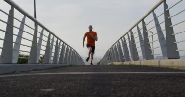 Genç bir atletik beyaz adam bir şehirde yaya köprüsünde egzersiz yapıyor, ağır çekimde çok hızlı koşuyor. Sağlıklı kentsel aktif yaşam biçimi.