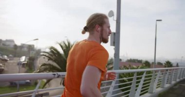 Şehirdeki bir yaya köprüsünde egzersiz yapan, kulaklıkları ağır çekimde müzik dinleyen genç bir beyaz adam. Sağlıklı kentsel aktif yaşam biçimi.