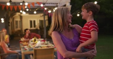 Beyaz tenli, son sınıf öğrencisi bir kadın torununu kucağında taşıyarak bahçede vakit geçiriyor. Ailesi arka planda, ağır çekimde gülümsüyor..