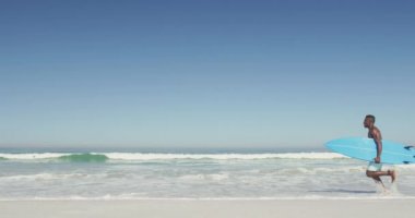 Afrika kökenli Amerikalı bir adamın tatilde tropikal bir plajda güneşlenirken mavi bir sörf tahtası taşıyarak ağır çekimde koşarkenki görüntüsü.