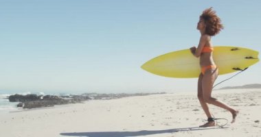 Afrika kökenli Amerikalı bir kadın, tropikal bir plajda güneşin tadını çıkarıyor, elinde sarı bir sörf tahtası, ağır çekimde denize doğru koşuyor. Plaj tatili eğlence aktiviteleri, sörf..