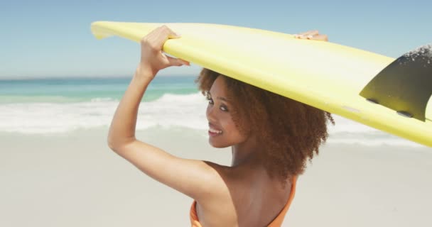 アフリカ系アメリカ人の女性の休日の太陽の下で熱帯のビーチに立って彼女の頭の上にサーフボードを保持し カメラになって笑顔 スローモーションで ビーチホリデーレジャーアクティビティ サーフィン — ストック動画