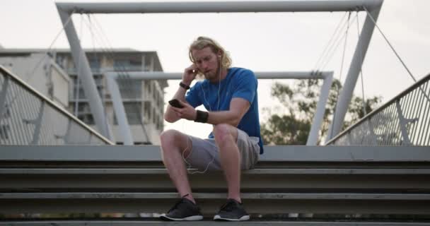 一个年轻的白人运动员在城市的人行天桥上锻炼 在慢动作的间歇中坐在台阶上使用智能手机 健康的城市生活方式 — 图库视频影像