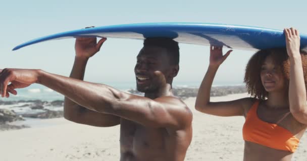 アフリカ系アメリカ人のカップルの休日の太陽の下で熱帯のビーチに一緒に頭の上にサーフボードを保持し 話をし 笑顔で スローモーションで歩く ビーチホリデーレジャーアクティビティ サーフィン — ストック動画