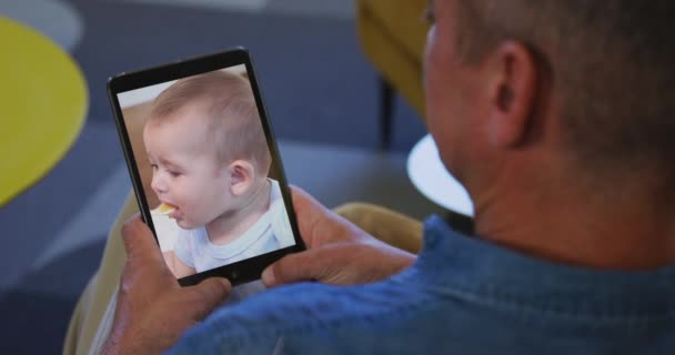 シニア白人男性に自宅でビデオチャット上のタブレットで男と赤ちゃん 一緒にオンラインで相互作用社会的距離と隔離されたロックダウンでの自己分離 — ストック動画