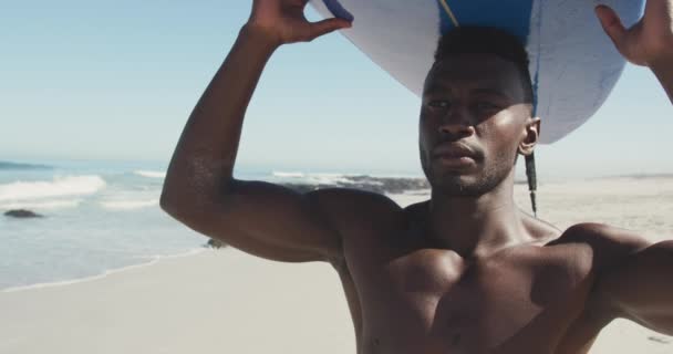 アフリカ系アメリカ人の男性は 太陽の下で熱帯のビーチに立っている彼の頭の上にサーフボードを保持し 道を探して笑顔で スローモーションで ビーチホリデーレジャーアクティビティ サーフィン — ストック動画
