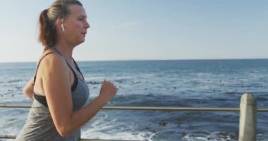 Deniz kenarındaki gezinti güvertesinde spor kıyafetleriyle çalışan, kulaklıkla ağır çekimde koşan son sınıf beyaz kadın. Emeklilik sağlıklı yaşam tarzı aktivitesi.