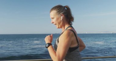 Deniz kenarındaki gezinti güvertesinde spor kıyafetleriyle çalışan, kulaklıkla ağır çekimde koşan son sınıf beyaz kadın. Emeklilik sağlıklı yaşam tarzı aktivitesi.