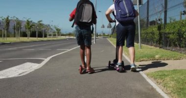Kafkasyalı ve Afrikalı Amerikalı çocuklar kaldırımda scooter sürüp okula giderken okul çantalarını yavaş çekimde taşıyorlar. İlkokul çocukları, yol güvenliği.
