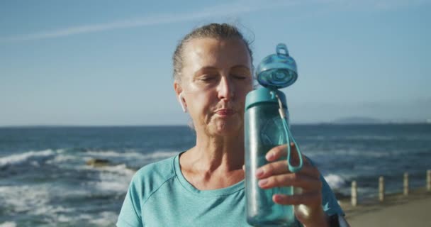 高个子白人妇女穿着运动服在海边散步 慢吞吞地从水瓶耳机里喝水 退休后健康生活方式活动 — 图库视频影像