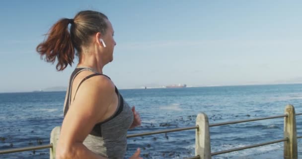 スポーツウェアを着て海沿いの遊歩道で働くシニア白人女性は ゆっくりとした動きでイヤホンで走っています 退職健康的なライフスタイル活動 — ストック動画