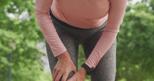 スポーツウェアを着て公園で働くシニアフィット白人女性は ゆっくりとした動きでイヤホンで負傷した膝を保持します 退職健康的なライフスタイル活動 — ストック動画